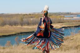 A Indigenous dancer in brightly coloured Fancy Shawl regalia beside a river.