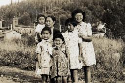 Black-and-white image of women and children standing on a gravel road in front of a field.