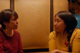 Two Inuit women sit in a dim room facing one another in intimate conversation. One is wearing a red sweater, the other a yellow sweater.