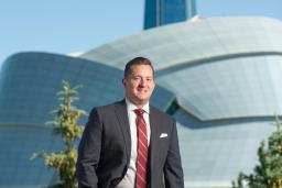 A person standing with the Museum in the background. He is wearing a suit, smiling, with his right hand in his pocket, looking directly at the viewer.