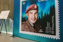 An enlarged Canadian stamp showcases a man with a red military beret with an aurora borealis and darkened forest in the background.