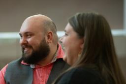 A man and woman sit together at a dinner table, smiling warmly as they listen to other guests. The man wears a salmon shirt with a gray vest, and there are small candles and drinks on the table, creating a cozy intimate atmosphere.