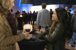 Two smiling women drink wine in a large room full of people.