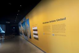 A yellow wall with the words "Ukrainian Artists United" and a dynamic blue and yellow graphic is right side of the image. On the left side, an image is projected on a screen showing a person with headphones and an open mouth, as though they are singing. Benches for sitting are placed in front of the screen.