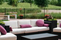 Couches and chairs sit in a fenced patio. Small trees and tall green prairie grass surrounds the patio. In the background, the spire from Esplanade Riel rises above the trees.