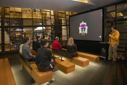 A small group of people sitting on wooden benches in a theatre face a screen showing an older woman seated in a chair. A man in a yellow shirt is standing to the right of the screen holding a microphone.