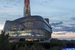 Exterior photo of the Canadian Museum for Human Rights’ glass windows and tower, taken against a prairie sky at dusk. The Winnipeg sign at the Forks is situated on the right side of the image. Multiple office towers and downtown lights are on the left-hand side of image.