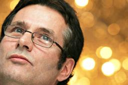 Close-up of a man’s face looking skyward in a thoughtful pose. He is wearing glasses and there are blurry spots of light in the background.