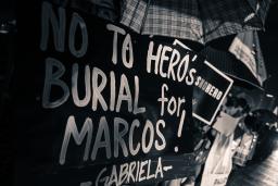 People standing in the rain under umbrellas along a roadside. Headlights from vehicles passing them are on the right side of the image. In the left foreground a sign blocks their faces from view and reads “No to hero’s burial for Marcos! Gabriela.”
