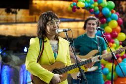 Two guitar players in bright and colourful outfits sing into microphones in front of a balloon arch.