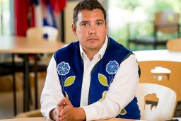 A man wearing a blue vest with flower beadwork, seated with his hands resting on a table.