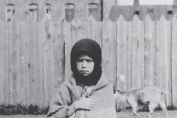 A young girl stands facing the camera with a sorrowful expression, clutching a shawl around her.
