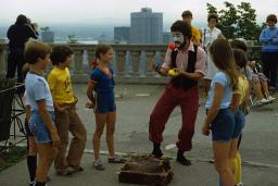 A man with a painted face juggles outdoors, as smiling children watch. A city skyline appears behind him at a distance.