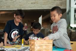 A child reaches into a basket of craft supplies while others work on their projects at a table.