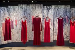 Five red dresses, representing missing and murdered Indigenous women and girls, hang in a row in the Canadian Journeys gallery as part of an exhibit titled “From Sorrow to Strength.” Behind the dresses, the scene of a snow-covered forest with more red dresses hung on the branches of trees is displayed on a series of banners.