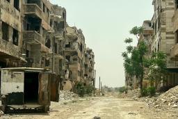 An abandoned city street where the building windows are gone, rubble lies piled in the road and a truck sits abandoned with its back doors open.