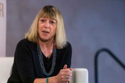 A woman sitting on a chair with hands clasped looks thoughtfully ahead, as if answering a question.
