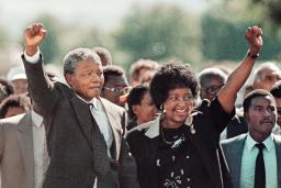 Nelson Mandela, an older black man with graying hair, forms a raised fist with his right hand and holds it above his head. With his left, he is holding hands with Winnie Mandela, a black woman with dark hair. She is smiling and has formed a raised fist with her left hand, mirroring Nelson Mandela’s raised fist. A large crowd is gathered behind them.