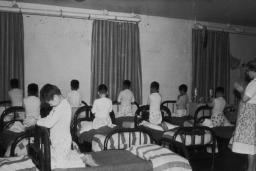A group of boys wearing pyjamas kneels on single beds with heads bowed and hands clasped as if in prayer. A woman stands in the room with her hands clasped in a similar manner.
