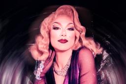 Drag queen with shoulder-length blonde hair wearing silver jewelry and a purple dress poses in front of a silver background.