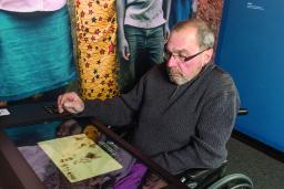 An older man sits in a wheelchair in front of a counter with a large touchscreen.