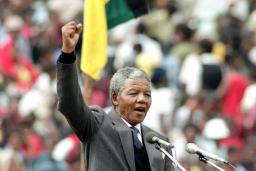 Nelson Mandela, an older dark-skinned man with short grey hair, stands in front of a podium and speaks into two microphones. He is wearing a grey suit with a tie. He is raising his right arm above his head and his hand is making a fist.