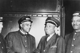 A black and white photo of four men in train porter uniforms. All of the men are smiling, and the two men in the middle appear to be shaking hands.