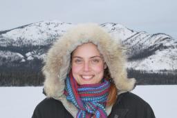 Smiling female wearing a fur-lined parka posing in front of a mountainous terrain.