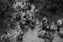 Rohingya women, children and men wade through waist-high muddy river water. Some carry young children, while others carry bags of possessions, including household items.
