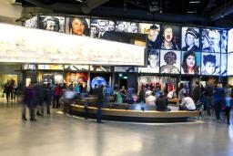 A large crowd in a museum gallery. There is a curved bench in the centre of the room. The display consists of a series of portraits above alcoves each containing a different exhibit.