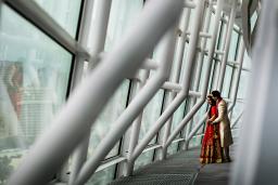 A man embraces a woman from behind as they stand together looking out of a wall of windows held together by a large white steel structure.