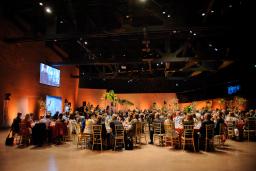 A crowd of people are seated around tables listening to a speaker in a large room.