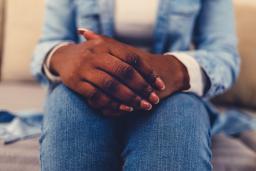 Close-up of a dark-skinned person sitting on a couch, with their hands clasped on their lap.