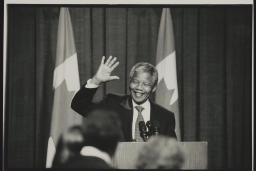 A black and white photograph of Nelson Mandela standing in front of a podium. He is smiling and waving.