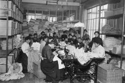 A large group of women sitting and sewing. Others are standing behind them in a room crowded with textiles. 