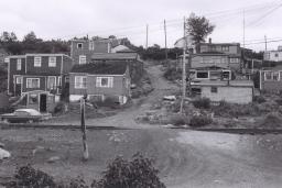 A black and white image of about a dozen houses on a hill. In the middle of the image, a dirt road travels up the hill between the houses.