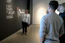 A woman points to a wall that says “Time to Act, Rohingya Voices” in both English and French while a small group of people stand and watch.