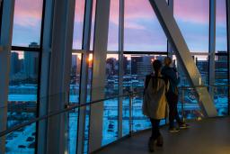 Two people gaze out a tall glass window at a sunset over a cityscape.