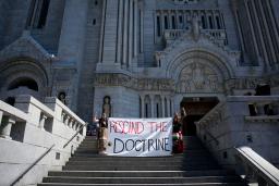Two people in braids and ribbon skirts raise fists and hold a large cloth banner reading “RESCIND THE DOCTRINE” on the steps of an enormous cathedral.