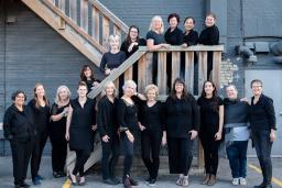 Women’s choir members are dressed in black and posed along an outdoor wooden staircase.