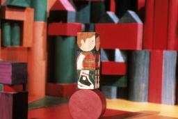 A wooden block painted with the image of a child, stands on top of a round block. Blocks of varying shapes, colours and sizes appear in the background.