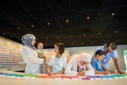 A family of six stands in front of curved table with coloured cards on it. Three family members are writing on cards with markers while the woman holds a smiling toddler. 