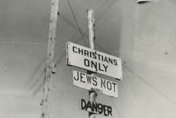 Black and white photo of three signs on a post, reading “Christians only,” “Jews not allowed” and “Danger.”