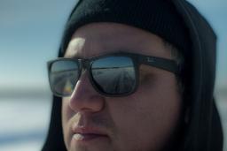 A close-up of a man wearing sunglasses and a hat, looking at something off camera. A road is seen reflected in his sunglasses.