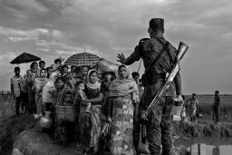 A soldier with a rifle on his back blocks a group of Rohingya before they cross a makeshift bridge. His left hand is raised, gesturing for them to stop.