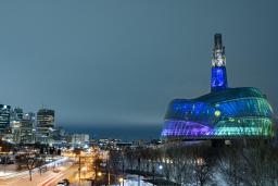 The CMHR’s exterior is seen at night with its glass panels lit up blue, green and purple. 