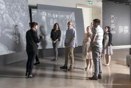 A small group of people stand in front of large screens displaying photographs and look at a woman who is speaking to them.