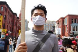 A man wearing a cloth mask and holding a sign, walking with a group of protestors carrying signs about ending racist violence and anti-Asian racism.