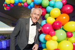 A performer wearing a tuxedo stands in front of a display of multi-coloured balloons.