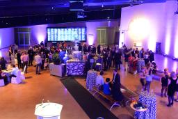 A party is taking place in a large room. Guests are gathered around tables holding glasses and chatting. Purple and blue light is cast throughout the room.